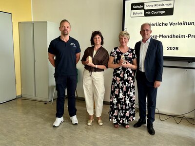 Zwei M&auml;nner und zwei Frauen posieren vor einem Beamer in einem Schulraum anl&auml;sslich einer Preisverleihung. 