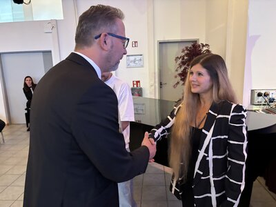Ein Mann im schwarzen Anzug reicht einer jungen Frau mit langen, dunkelblonden Haaren die Hand zur Gratulation. 