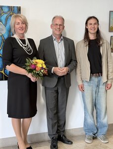 Eine Frau und zwei M&auml;nner vor einer Wand mit Bildern. 