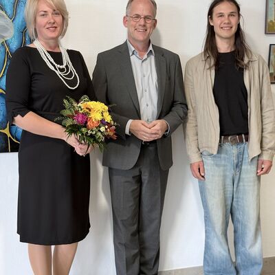 Eine Frau und zwei M&auml;nner vor einer Wand mit Bildern.